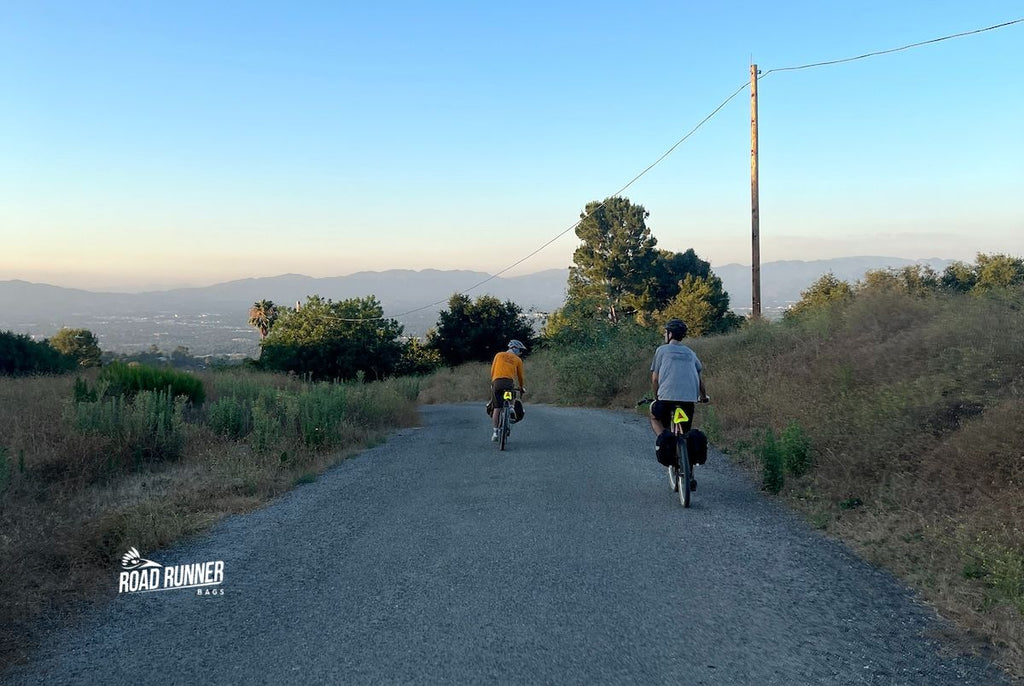 Two cyclists on gravel bikes loaded with Road Runner Bags on a dirt road west of Los Angeles are seen riding into the setting light