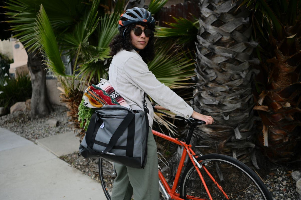 woman with dark curly hair wearing sunglasses, bike helmet, white shirt, green pants, and grey tote bag standing next to a bike on sidewalk. palm trees in background